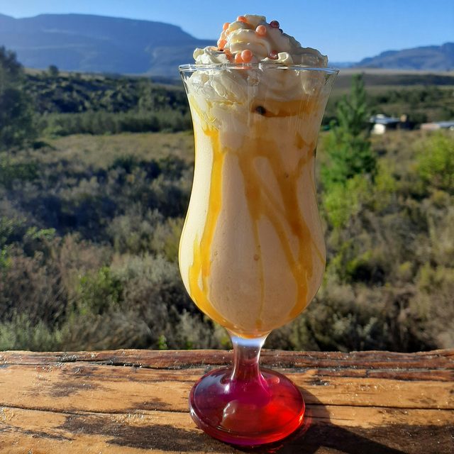 A caramel milkshake on a wooden railing with the veld and mountains behind